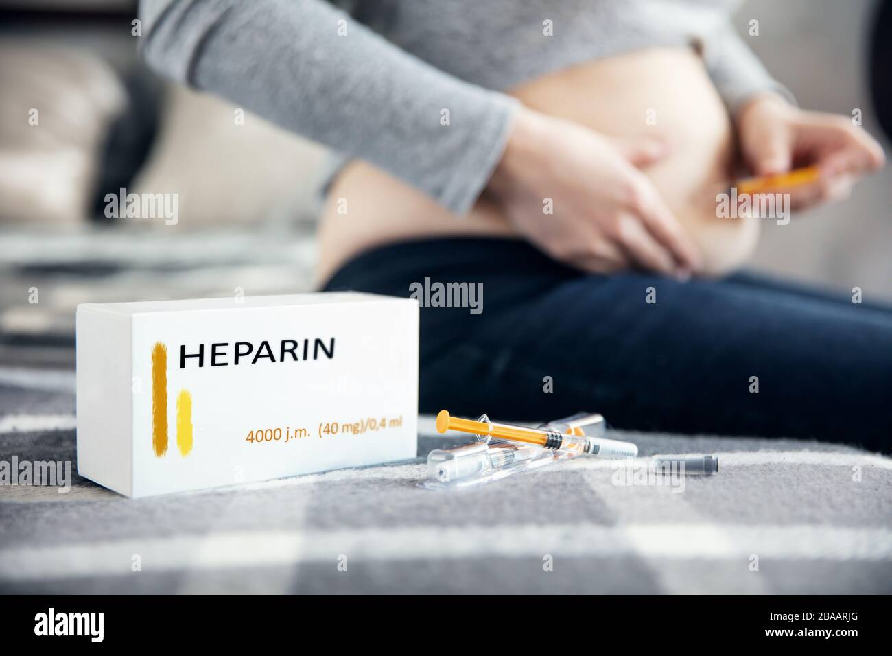 Syringes with medicine and a box with 'heparin' inscription lies on the blanket. Pregnant woman makes an injection in the background. Inscriptions on Stock Photo