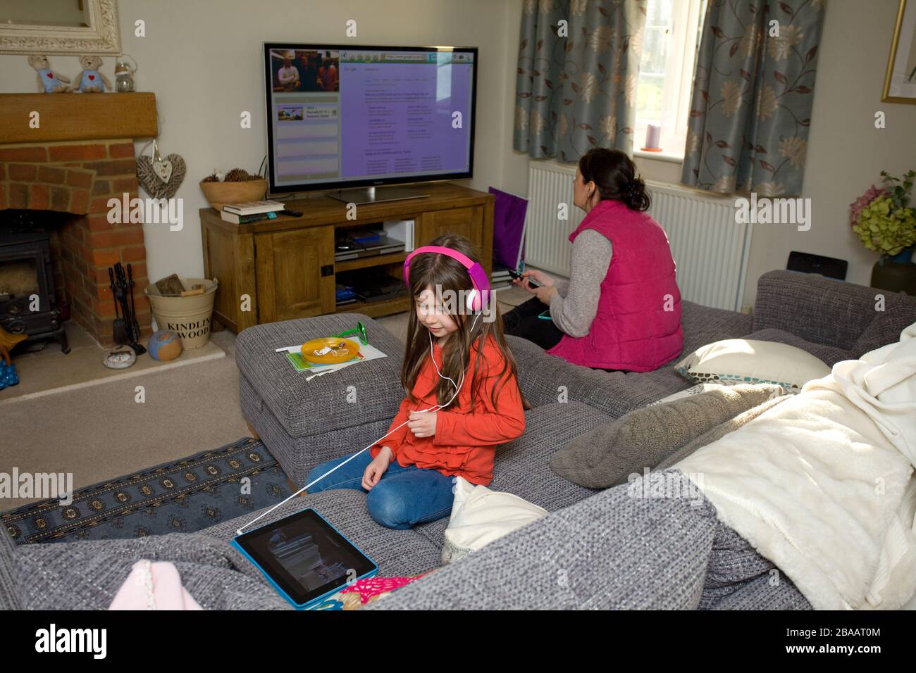 young girl wearing headphones watching iPad and her mother in the background watching television, England Stock Photo