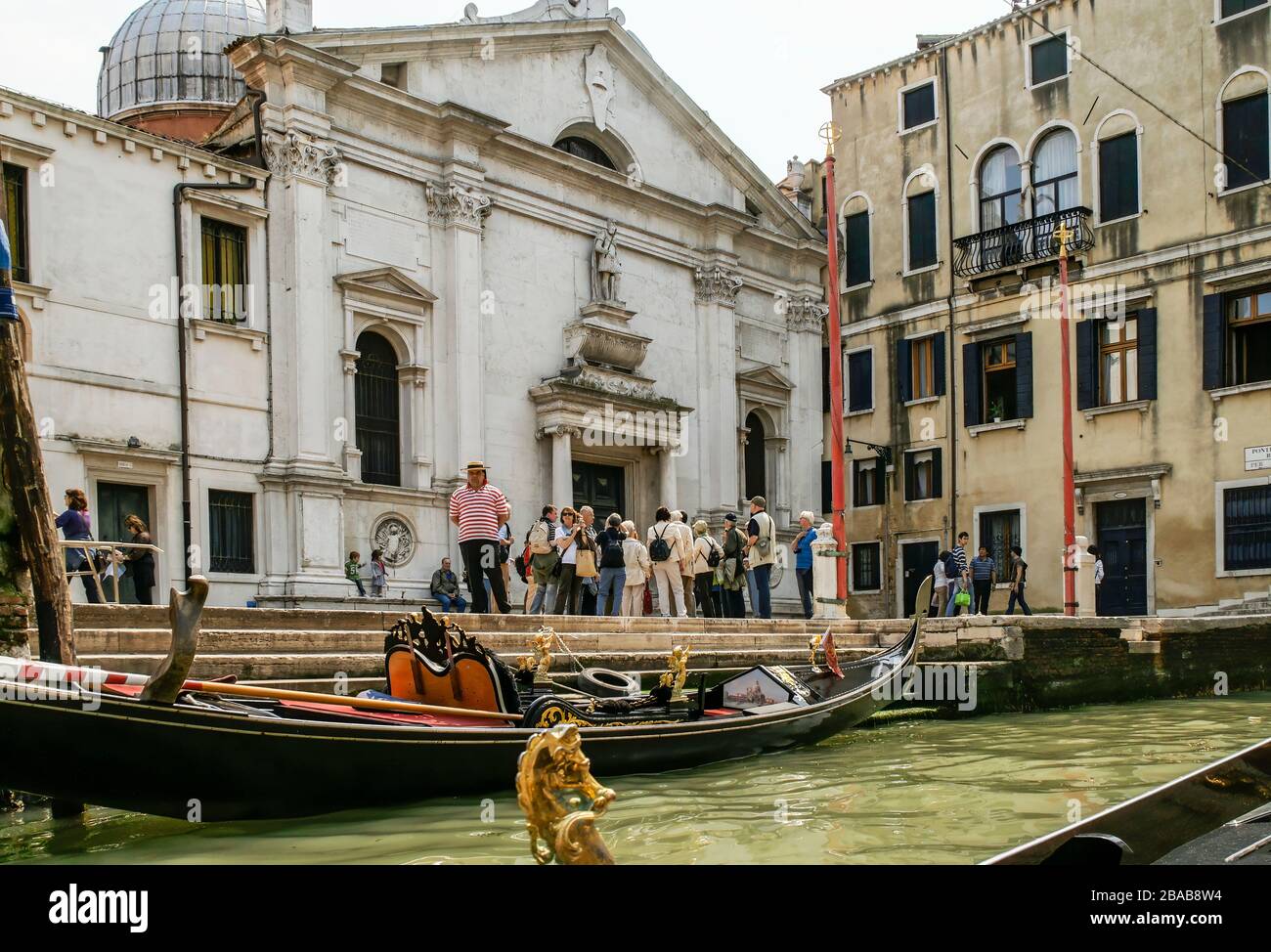 A gondola tied up in the canal with a gondolier waiting nearby. A tourist group gathers at the church. NMR Stock Photo