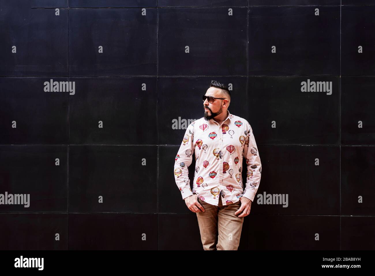Bearded man wearing sunglasses leaning on black wall, looking aside Stock Photo