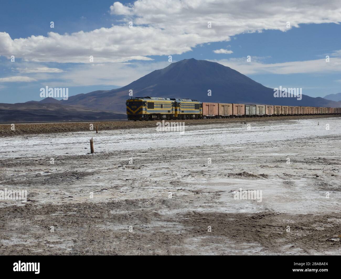 Long train with many container driving through remote area of bolivia close to salar de uyuni , with mountain , sky , salt ground looking like snow Stock Photo