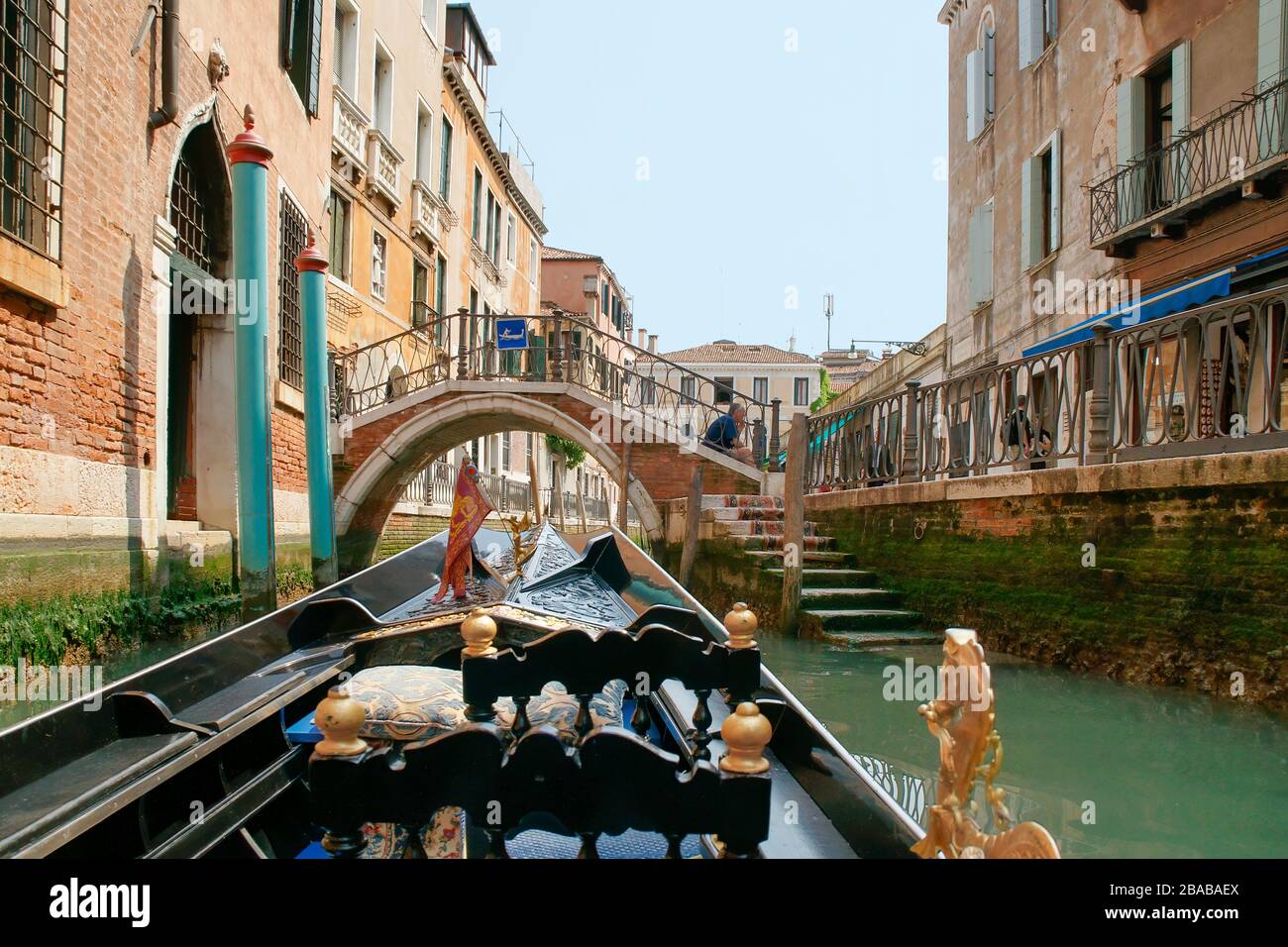 View while riding in a gondola down a small side canal with buildings and shops on the sides. A man sits on the steps of a small brick bridge. Venice, Stock Photo
