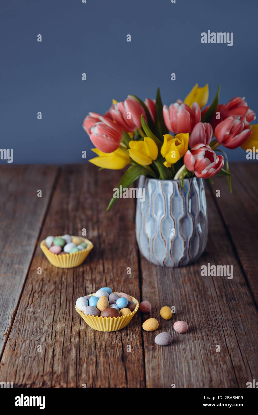 Easter candies covered eggs in various pastel colors in paper baskets near vase with fresh tulips on rustic wooden table and dark blue background. Fas Stock Photo