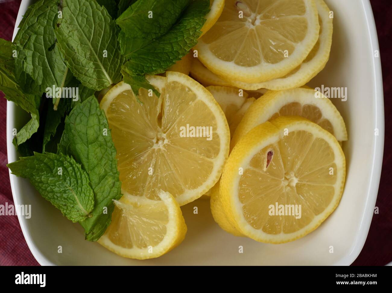 Fresh lemon slices, some with seeds, in a white bowl with mint leaves Stock Photo
