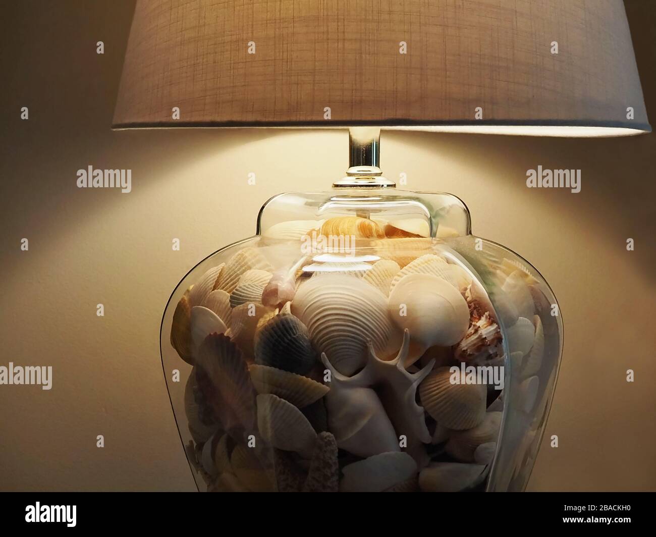 Closeup on a clear glass table lamp base filled with seashells, with a beige linen lampshade. Stock Photo
