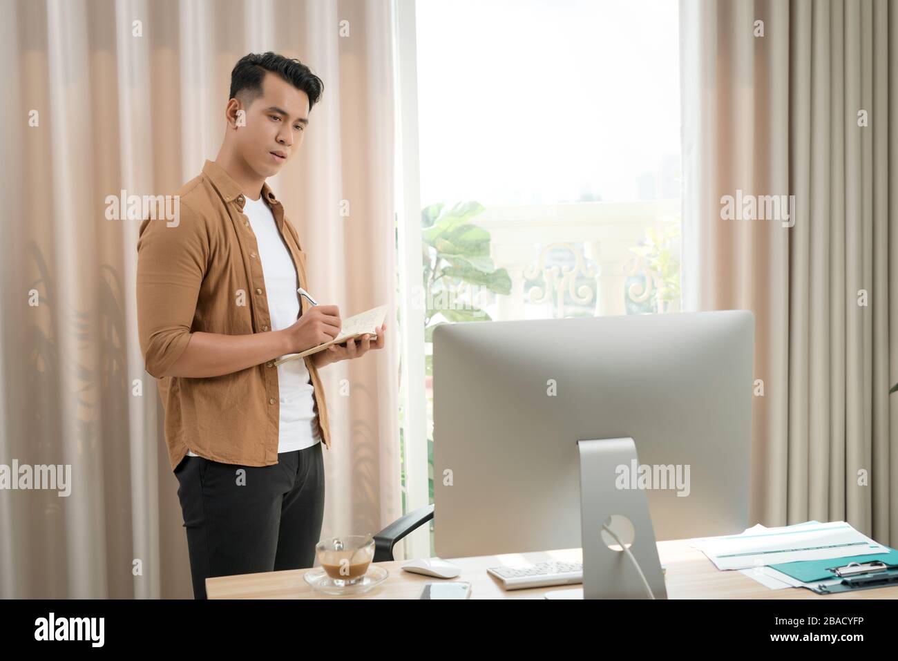 Asian man, aged 25-35 at the desk and smiling happily. He monitors sales on the computer screen. Stock Photo