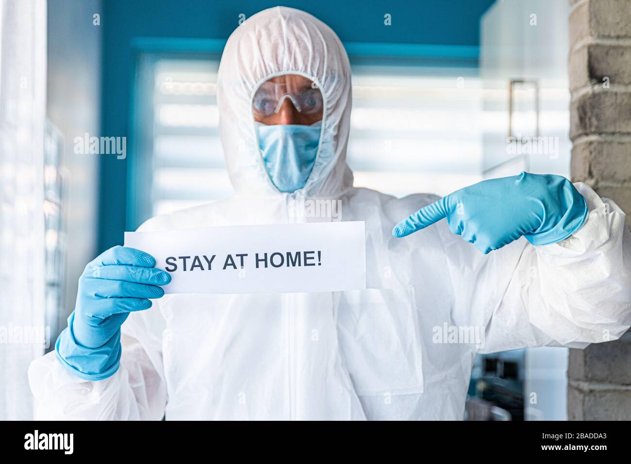 Man in protective anti virus suit and mask holding card with message stay at home Stock Photo