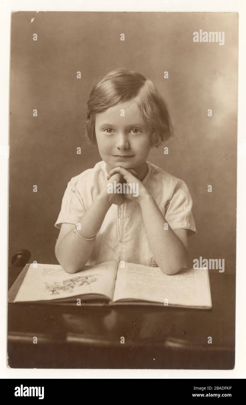 Early 1900's studio portrait of cute young girl with typical hairstyle, sitting with a book open, circa 1930's, U.K. Stock Photo