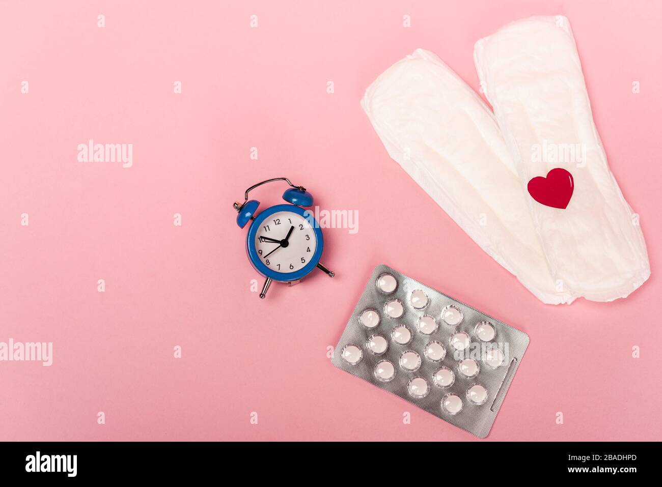 Top view of feminine pads with paper heart, birth control pills and alarm clock on pink background Stock Photo