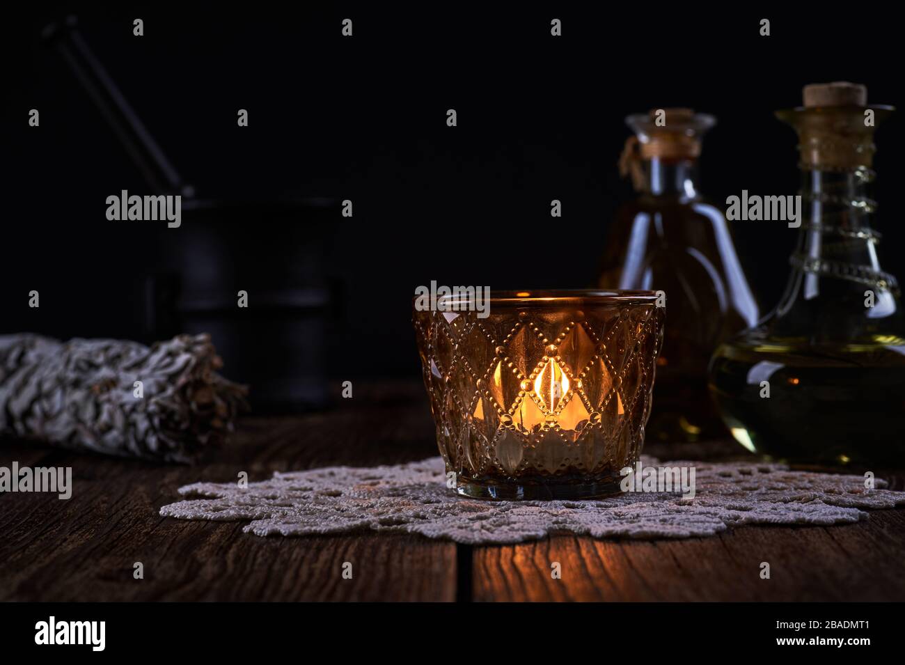 Esoteric still life with burning candle, green vials with drops of alternative medicine and iron mortar on blurred black background and old wooden tab Stock Photo
