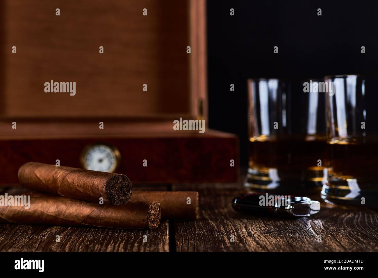 Still life with three cuban cigars, two glasses of whiskey or rum, lighter and wooden box with hygrometer. Old wooden table top and black background. Stock Photo