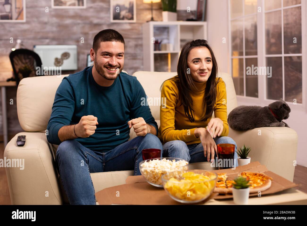 Happy couple cheering for their favourite team while watching TV in he living room. Stock Photo
