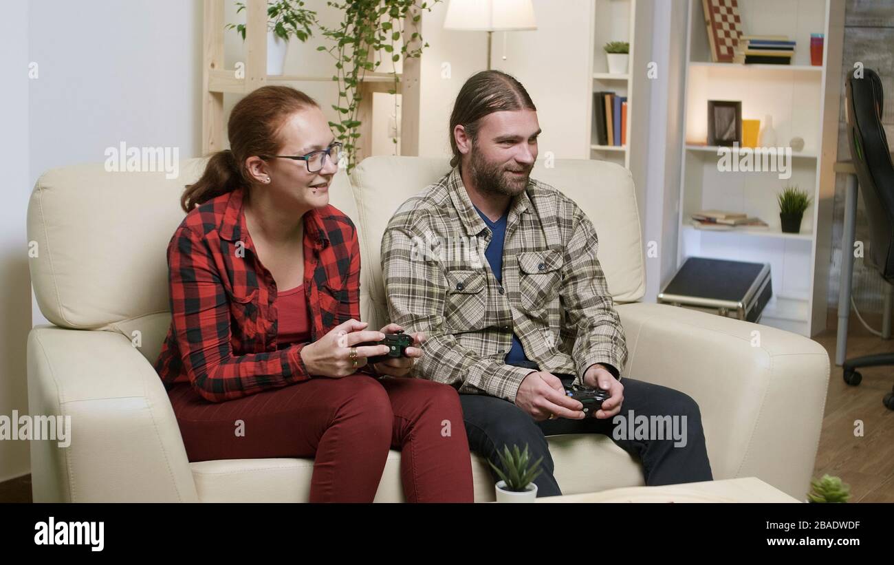 Husband and wife sitting on sofa playing video games using wireless controller. Stock Photo