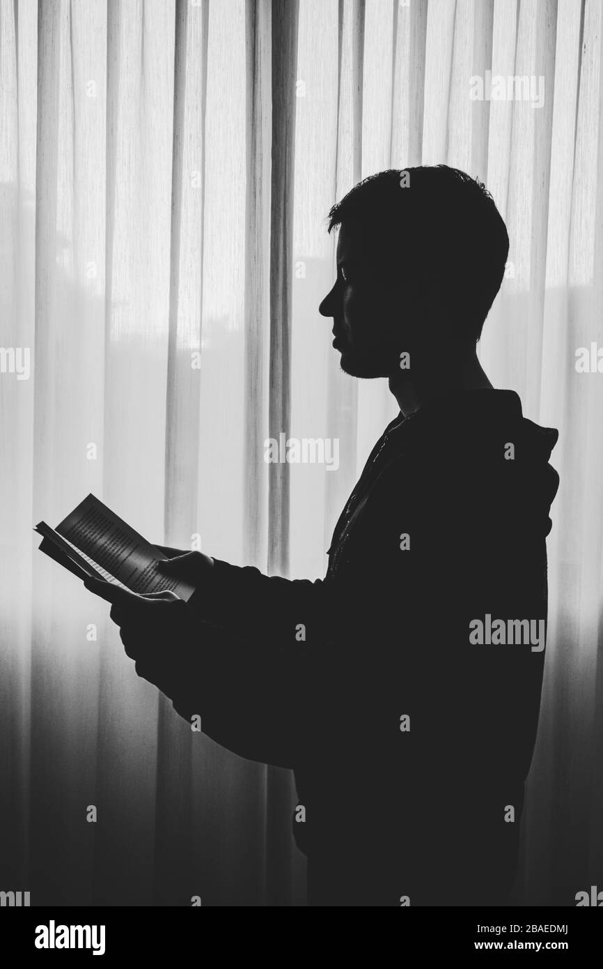 Side view of silhouette boy standing by window reading a book Stock Photo