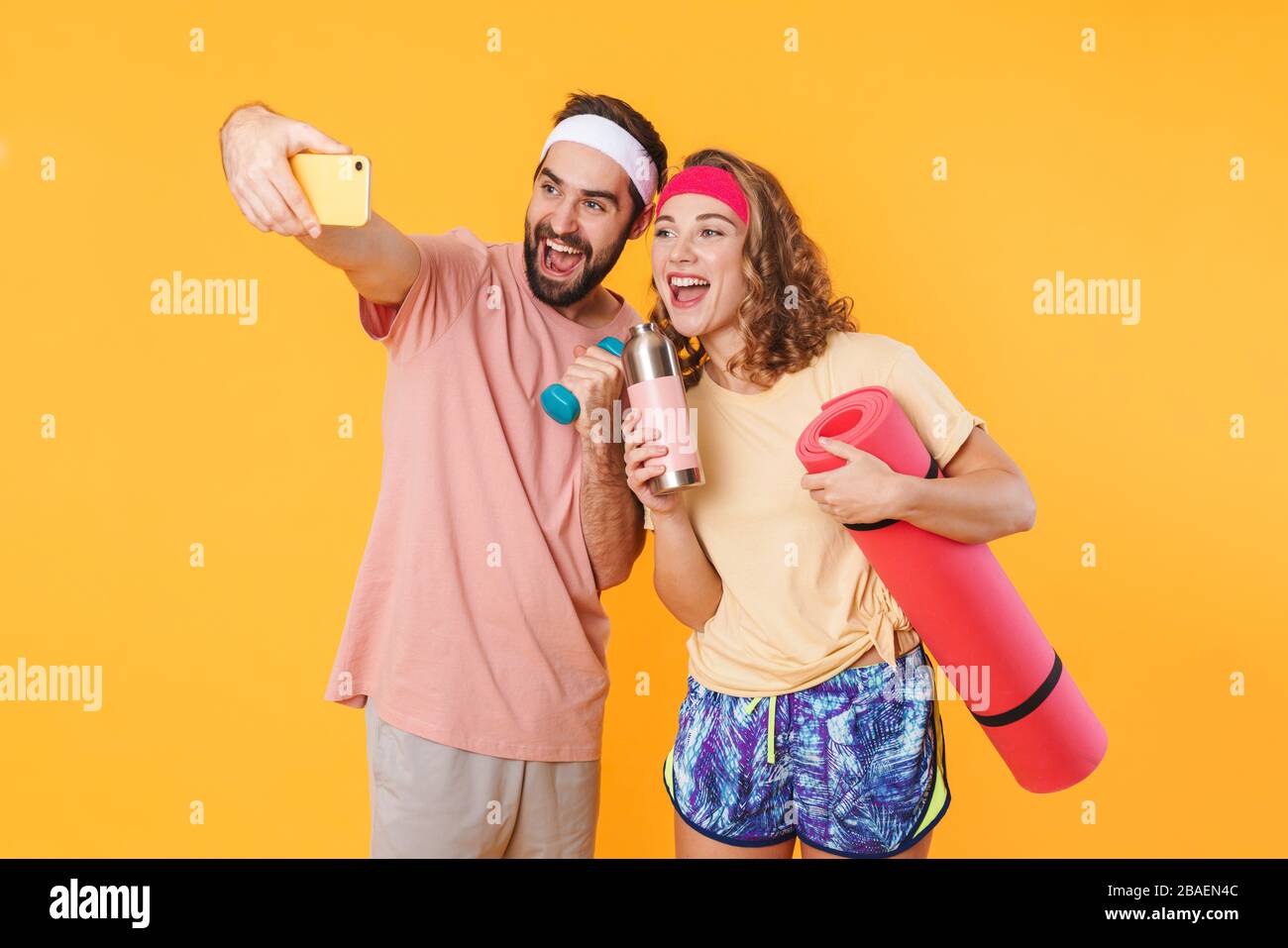 Portrait of athletic young happy couple taking selfie photo with dumbbells and fitness mat isolated over yellow background Stock Photo