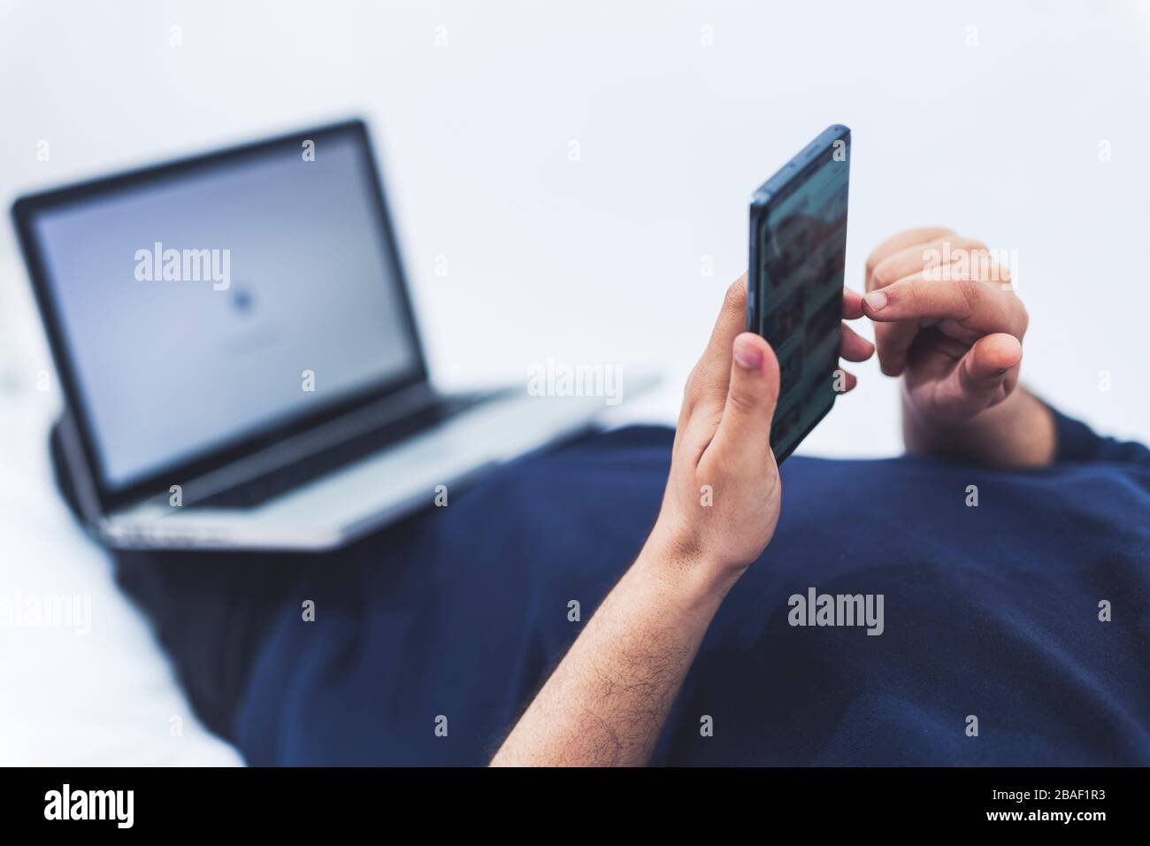 close up view of a man using his mobile phone and a lap top while lying on bed Stock Photo