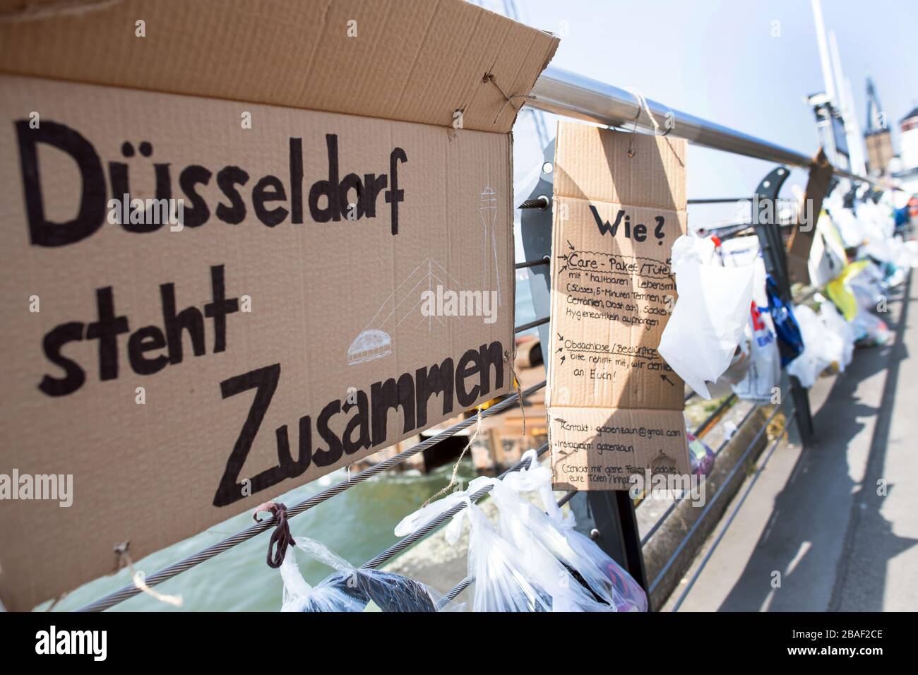 Duesseldorf, Germany. 27th Mar, 2020. Bags of food hang on a gift fence on  the banks of the Rhine, next to which hangs a cardboard sign "Düsseldorf  stands together". Homeless people in