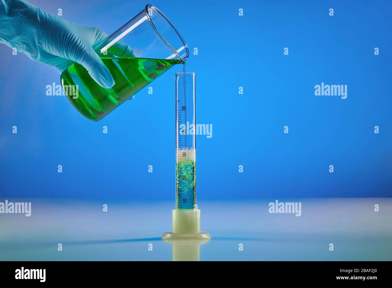Hand in disposable glove is pouring green liquid from beaker into measuring  cylinder, blue background. Laboratory research of Coronavirus. Worldwide p  Stock Photo - Alamy