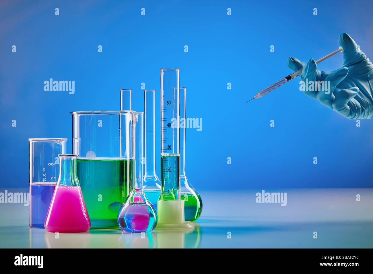 Hand in disposable glove holding syringe. Colorful chemical reagents in beakers, medical flasks and measuring cylinder, blue background. Coronavirus, Stock Photo