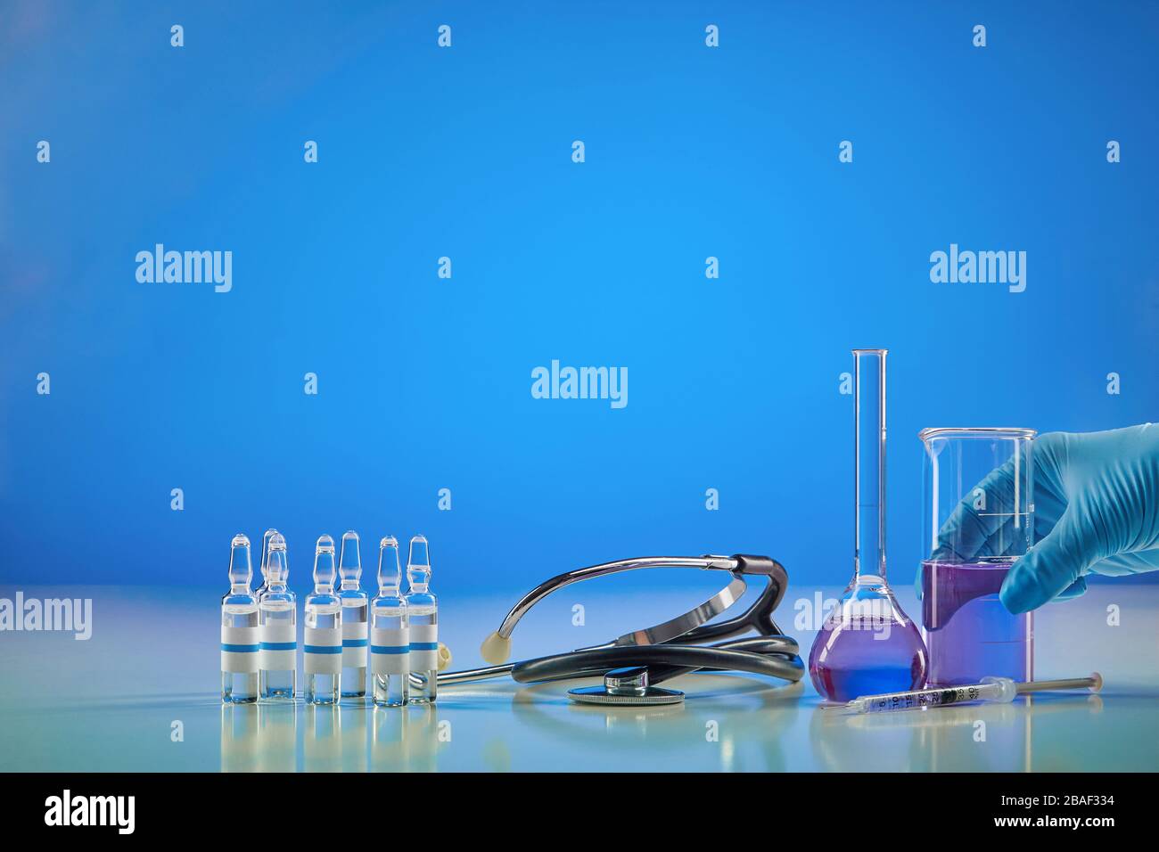 Hand in disposable glove holding beaker with purple chemical reagent. Six ampoules with liquid, syringe, medical flask and phonendoscope, blue backgro Stock Photo