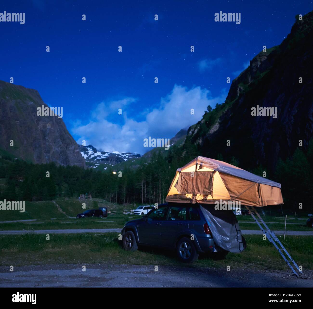 Camping night scene with silver car, majestic hills on background.  Beautiful view of blue evening sky over high mountains and SUV with rooftop  tent. Concept of travelling, camping and mountain hiking Stock