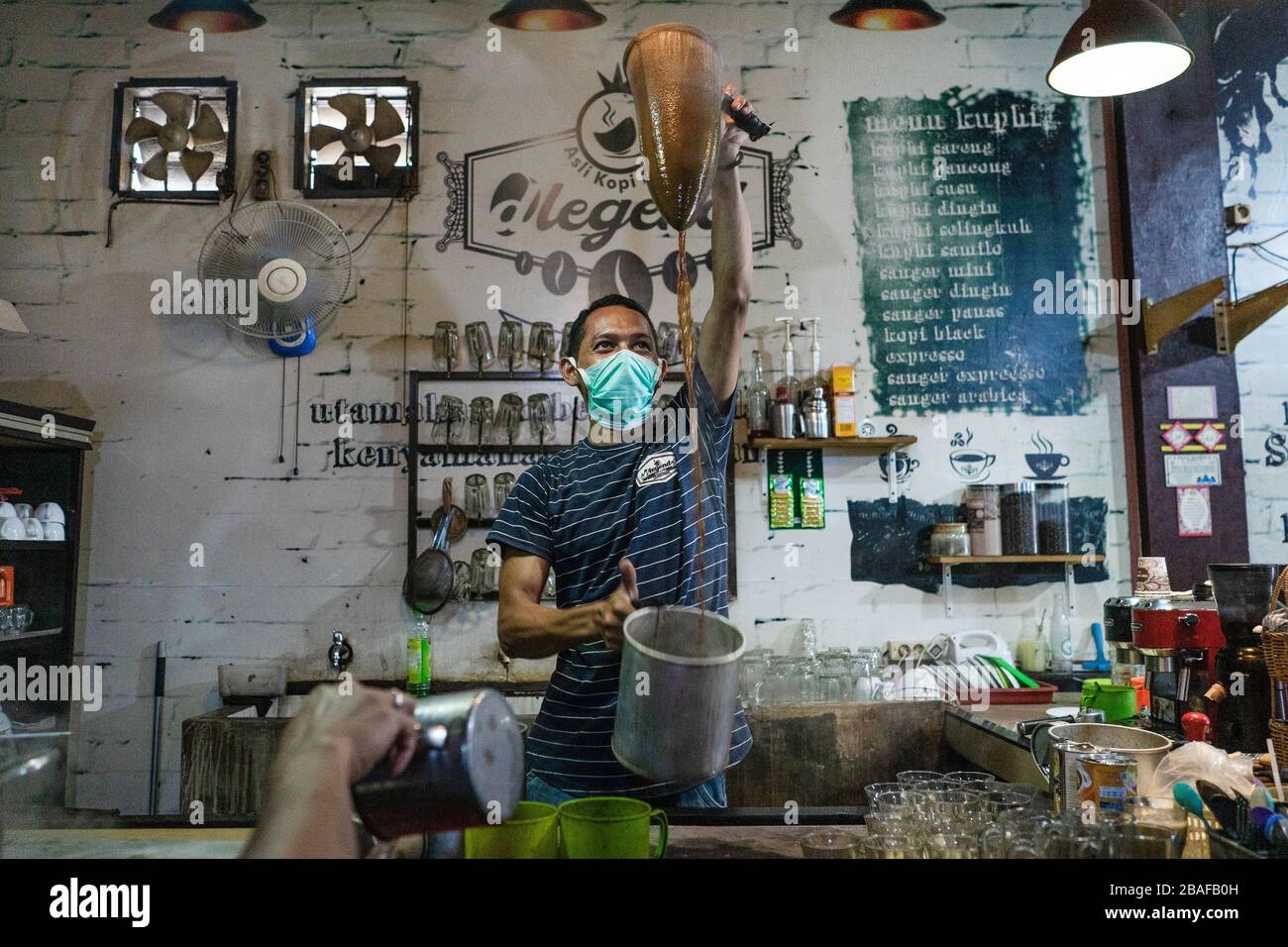 A barista wearing a face mask as a precaution against the spread of Coronavirus makes Acehnese coffee for his customers at a coffee shop in Lhokseumawe. Indonesian government on Friday, confirmed 1.046 for COVID-19 coronavirus cases, 46 people recovering and 87 deaths. Stock Photo