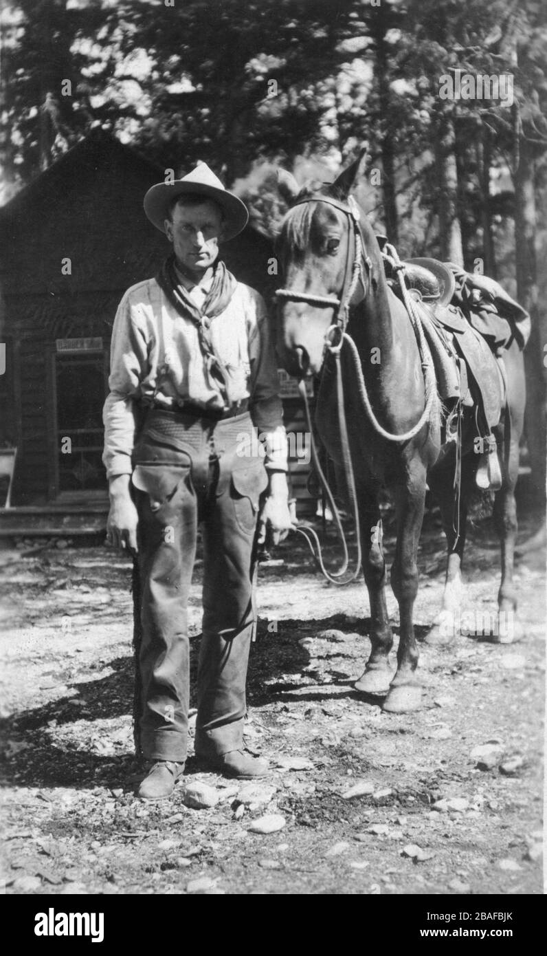Cowboy in America with his saddled horse. He is unarmed. The log cabin behind him has a sign that seems to say 'Barack     '  This might be a barracks building for cowboys or a camp or ??  An indicator that he might not be a real cowboy is that he is wearing shoes, not boots.   To see my Western-related  images, Search:  Prestor  vintage  west  vehicle Stock Photo