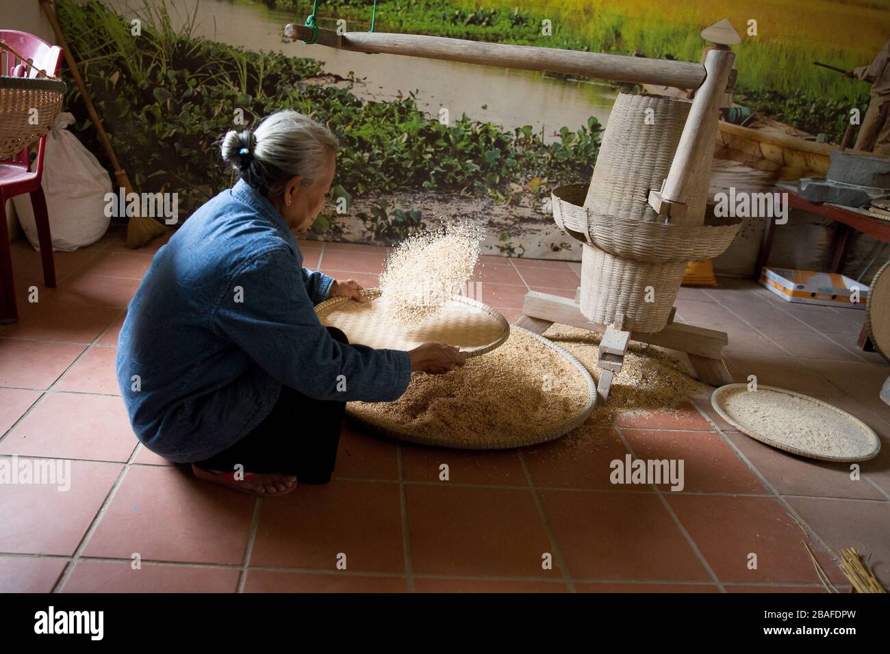 A Vietnamese woman crouches down to sift rice with a large bamboo sieve Stock Photo