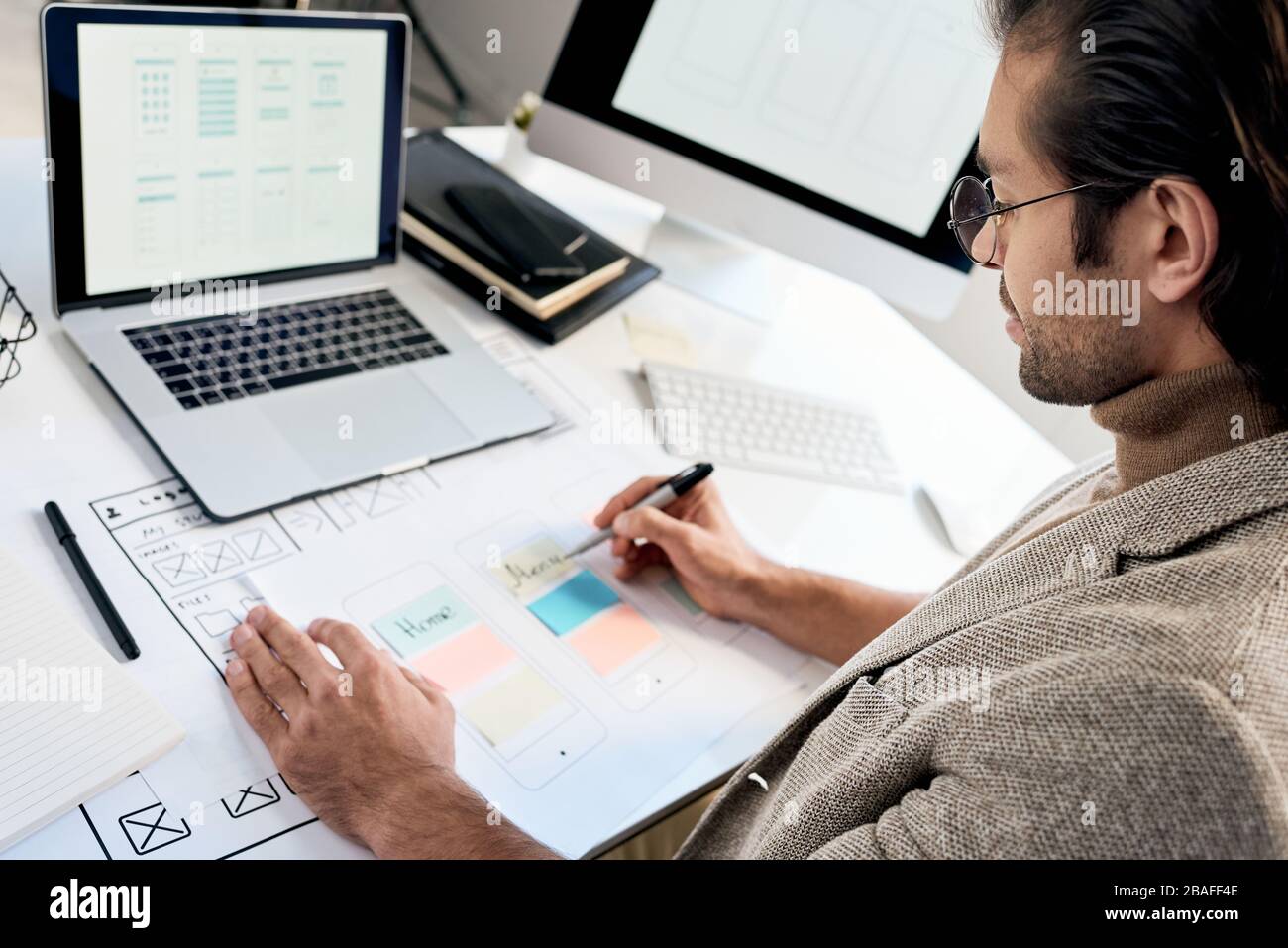 Serious phone interface engineer in glasses sitting at desk and making notes on interface sketch Stock Photo