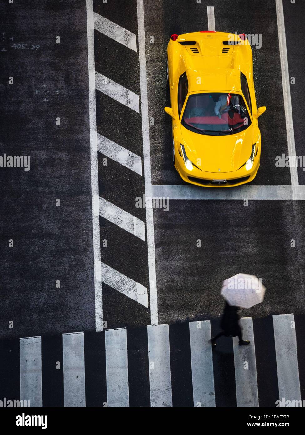 Tokyo Crossing in the Rain. View from above as a pedestrian with umbrella crosses in front of a waiting Ferrari. Stock Photo