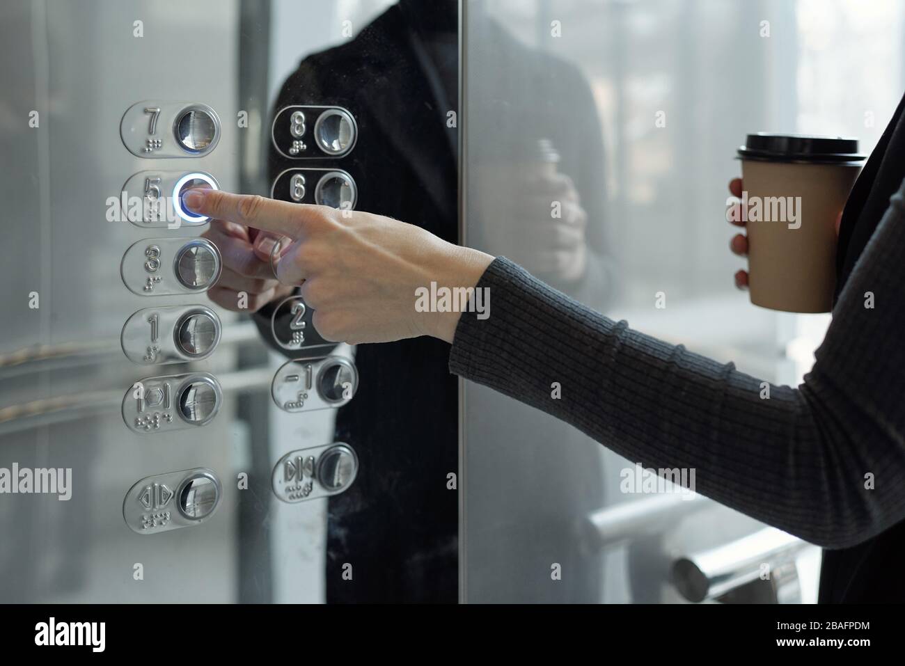Hand of young businesswoman with drink pushing button inside elevator while going back to office on fifth floor at coffee break Stock Photo