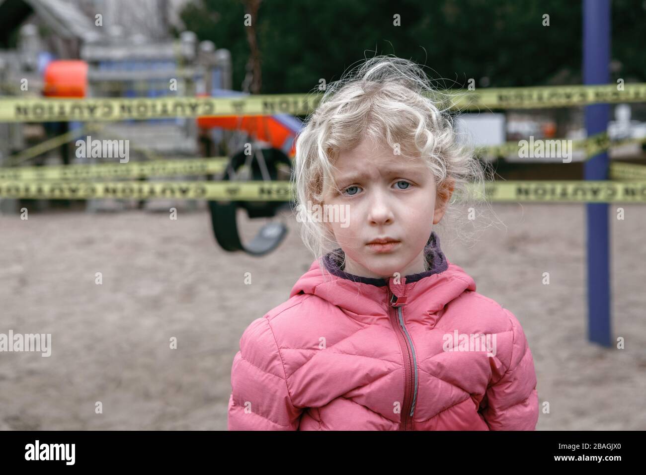 Portrait of sad upset Caucasian girl on closed playground outdoor. Kids play area locked with yellow caution tape in Toronto Canada. Coronavirus Stock Photo