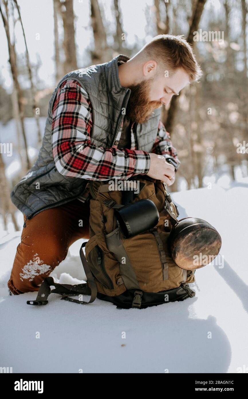man with bead wearing flannel and vest kneels in snow with pack Stock Photo