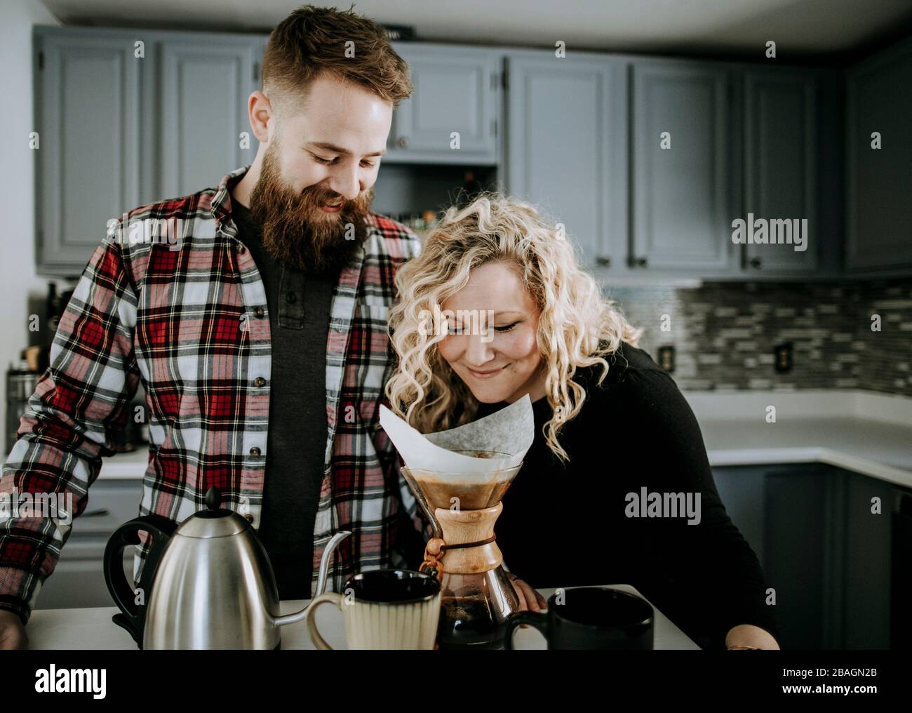 young female smells morning coffee while husband with beard looks on Stock Photo