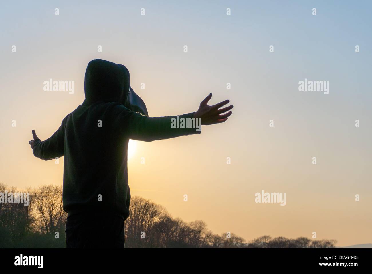 A hooded figure wearing a plague doctor mask, with arms out stretched. At sunset on a winters day. Stock Photo