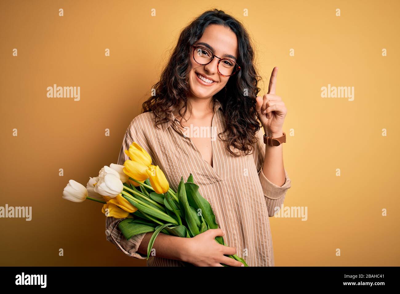 Young beautiful romantic woman with curly hair holding bouquet of yellow tulips smiling amazed and surprised and pointing up with fingers and raised a Stock Photo