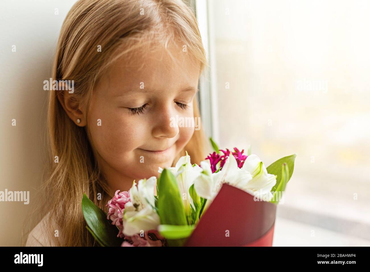 School kid in self isolation sitting near window and sniffing bouquet of flowers. Girl stay at home during covid-19 lock down, self isolation, stay Stock Photo