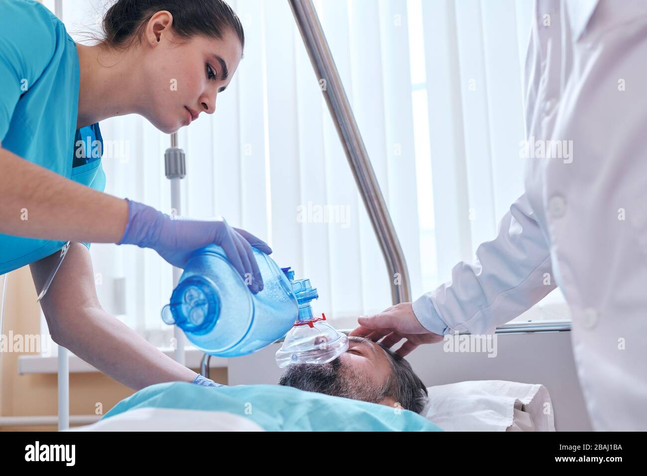 Young nurse in surgical gloves using bag valve mask while assisting ...