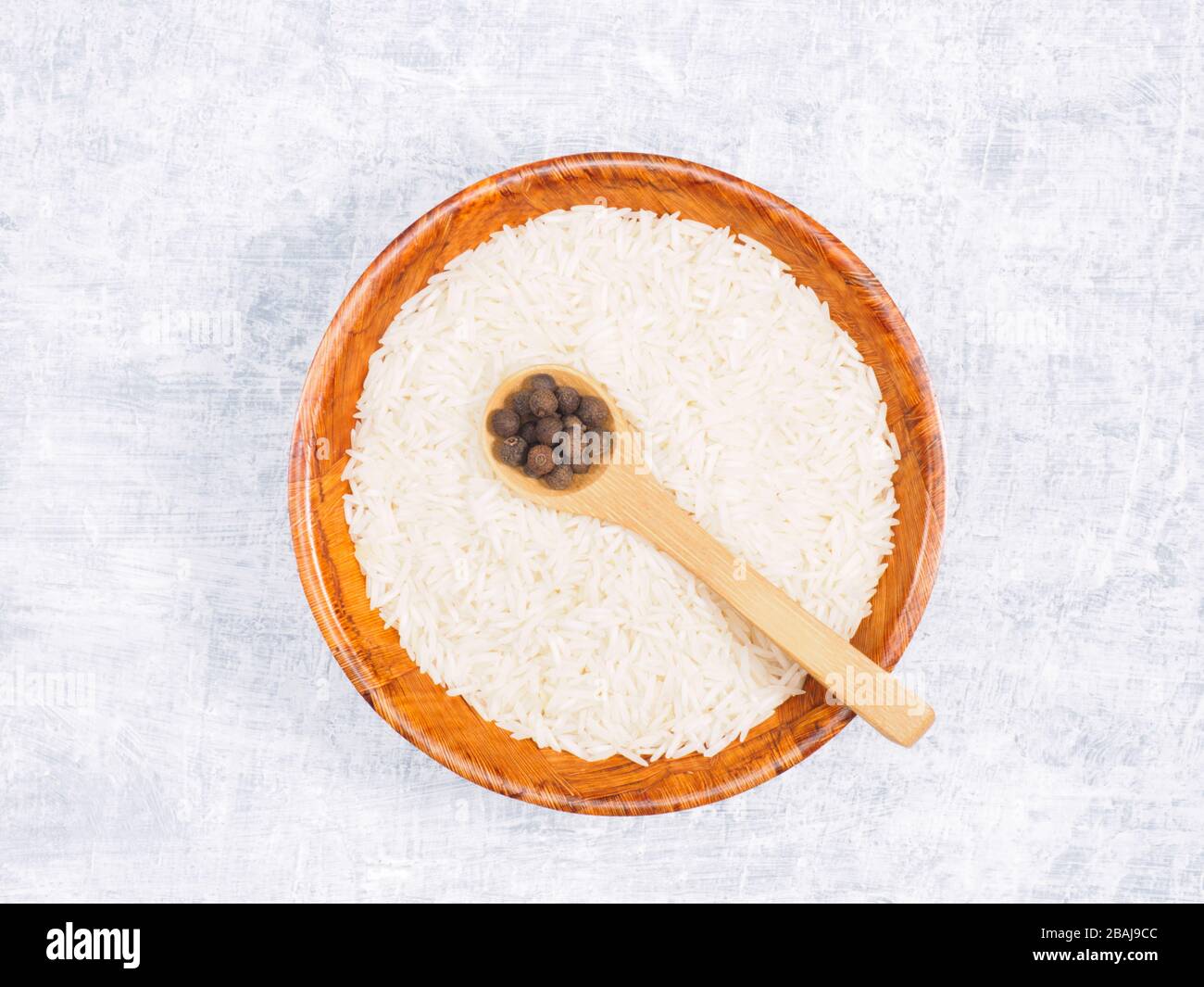 Indian long grain Basmati rice in brown wooden bowl and allspice in spoon on white concrete background Stock Photo