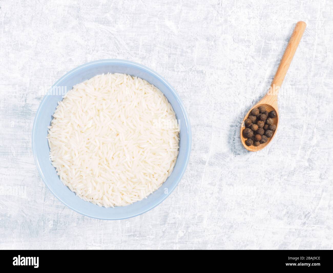 Indian long grain Basmati rice in blue ceramic bowl and allspice in spoon on white concrete background Stock Photo