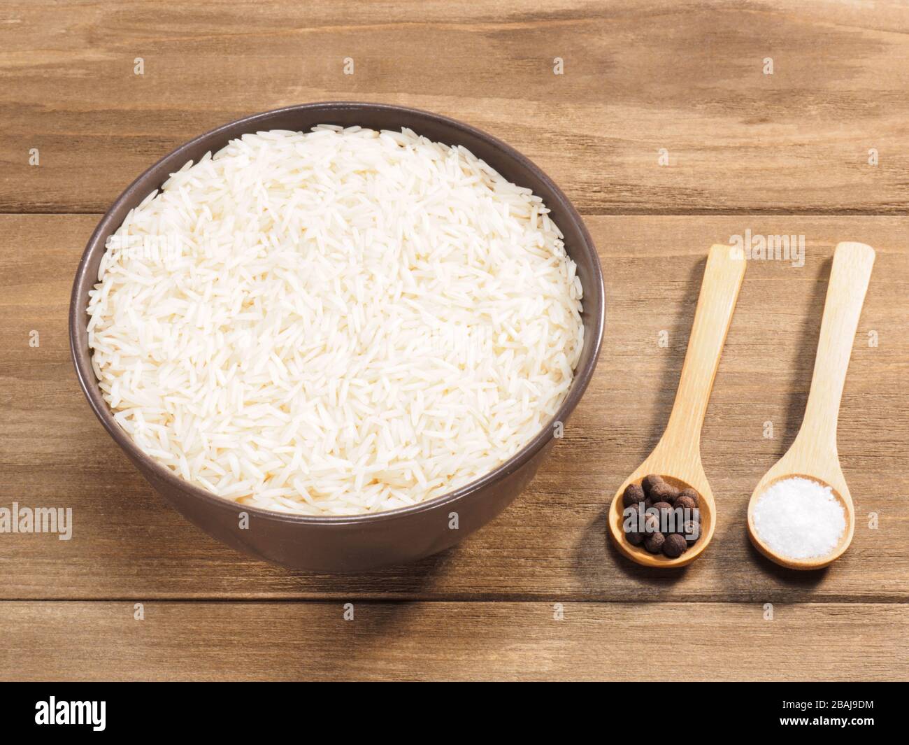 Long grain Basmati rice in brown ceramic bowl, allspice, salt on brown wooden background Stock Photo