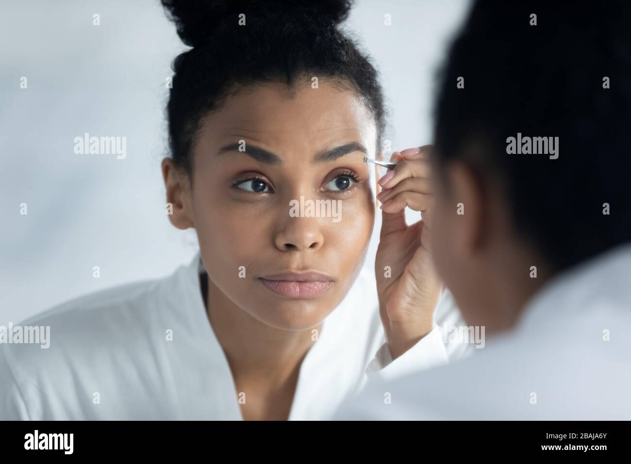 African woman looks in mirror plucking thick eyebrows using tweezers Stock Photo