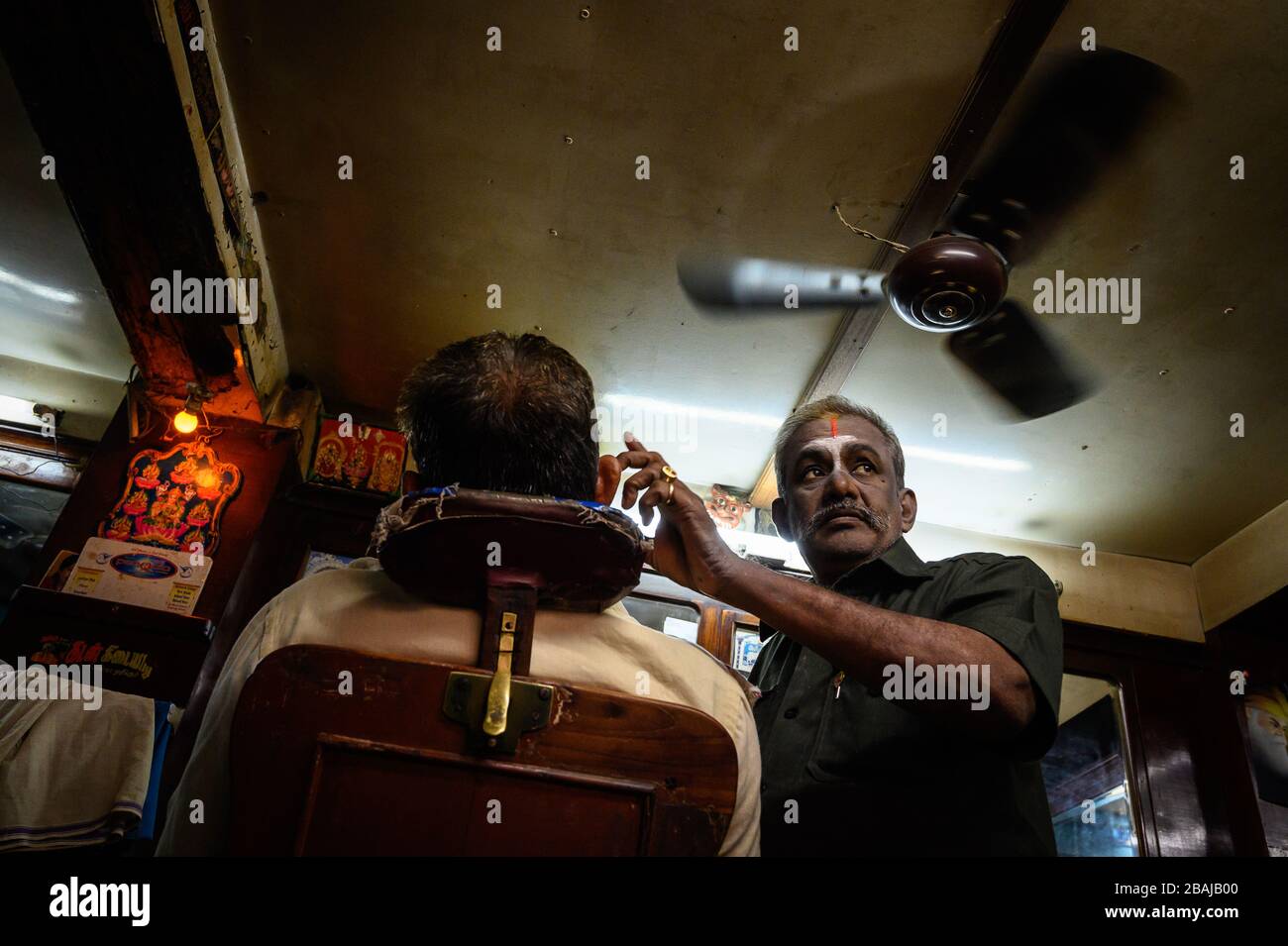 A barber tending to a customer under a fan in his shop , Madurai, India ...