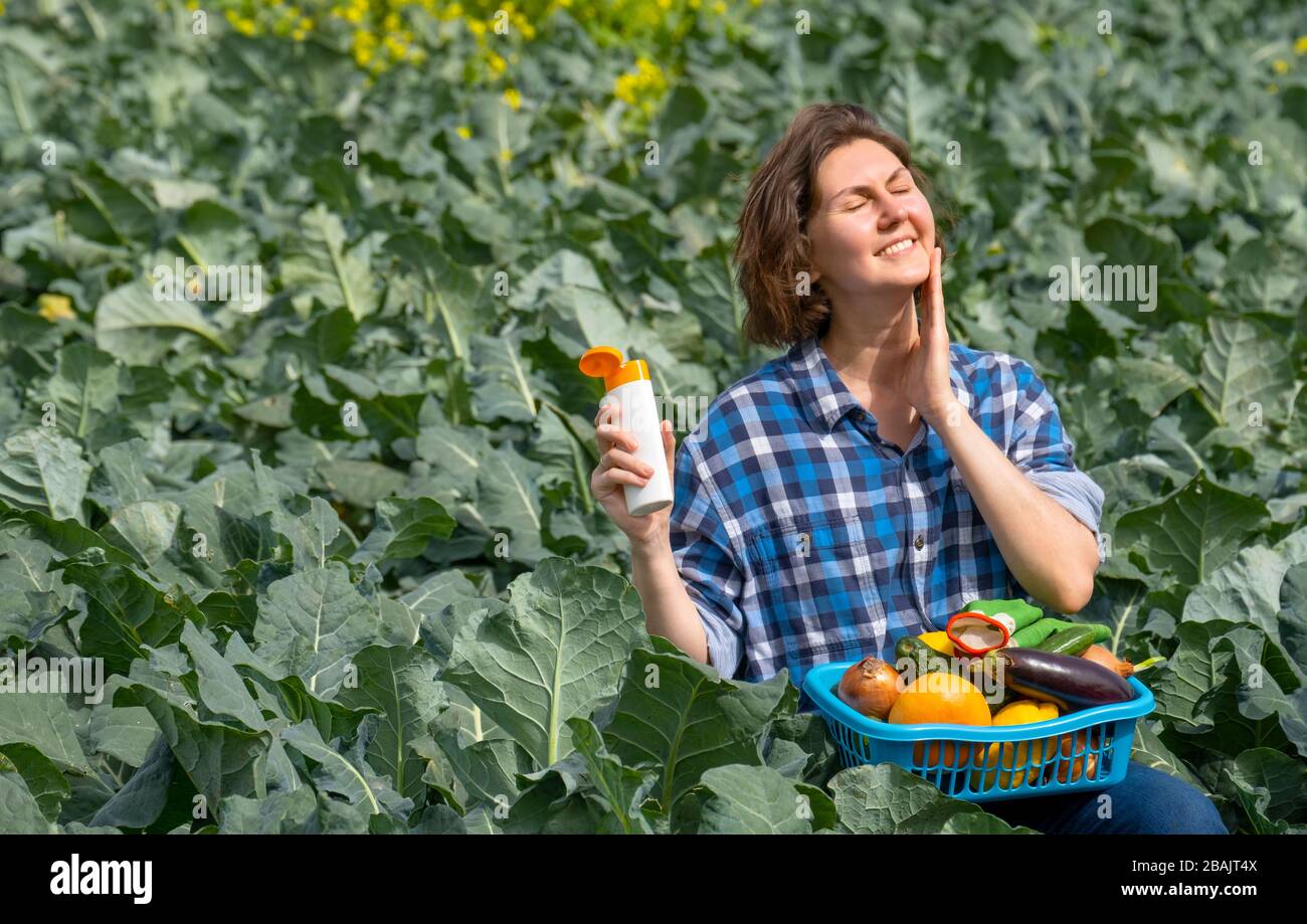 woman working on an agricultural field during a sunny day and protecting her skin from the sun with sunscreen. woman holds a basket with collected vegetables on her lap Stock Photo
