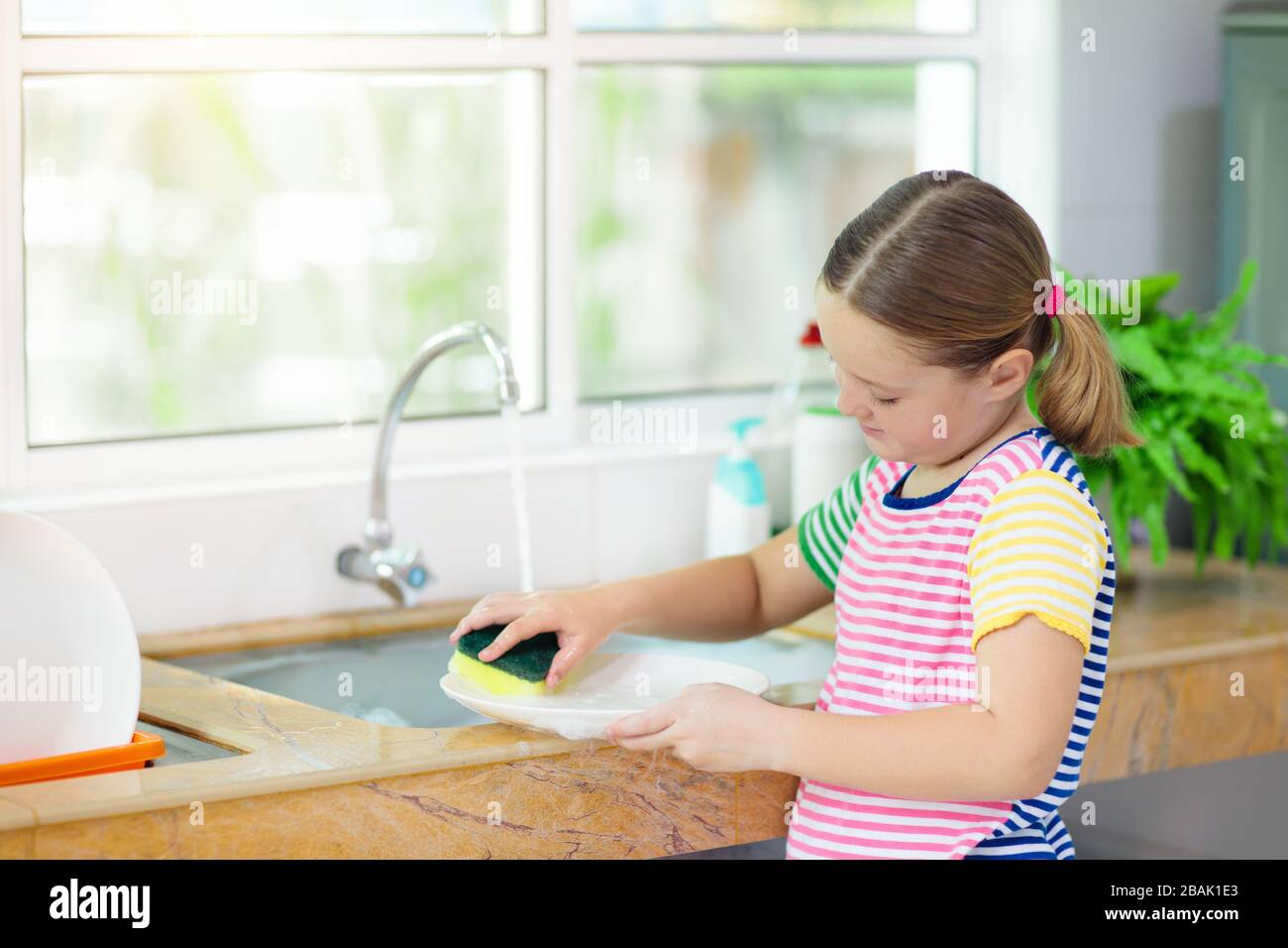 Child washing dishes. Home chores. Kid in white kitchen cleaning plates after lunch at window. Stock Photo
