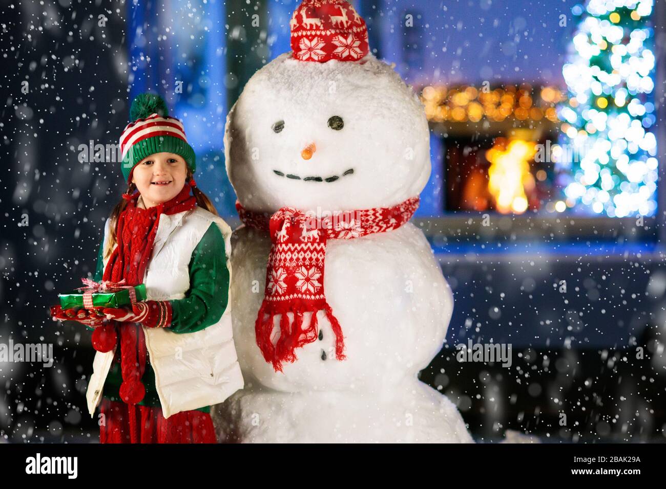 Kids build snowman on Christmas eve. Child with presents and gifts in snowy backyard next to window to living room with Christmas tree and decorated f Stock Photo