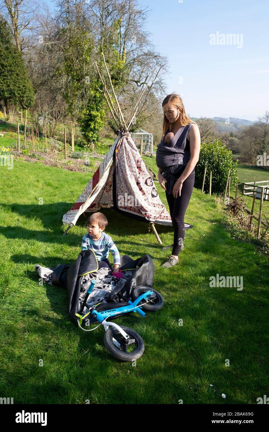 Mother with sling baby carrier and child boy children with  tipi playing in a country garden outside in spring 2020 Wales UK KATHY DEWITT Stock Photo