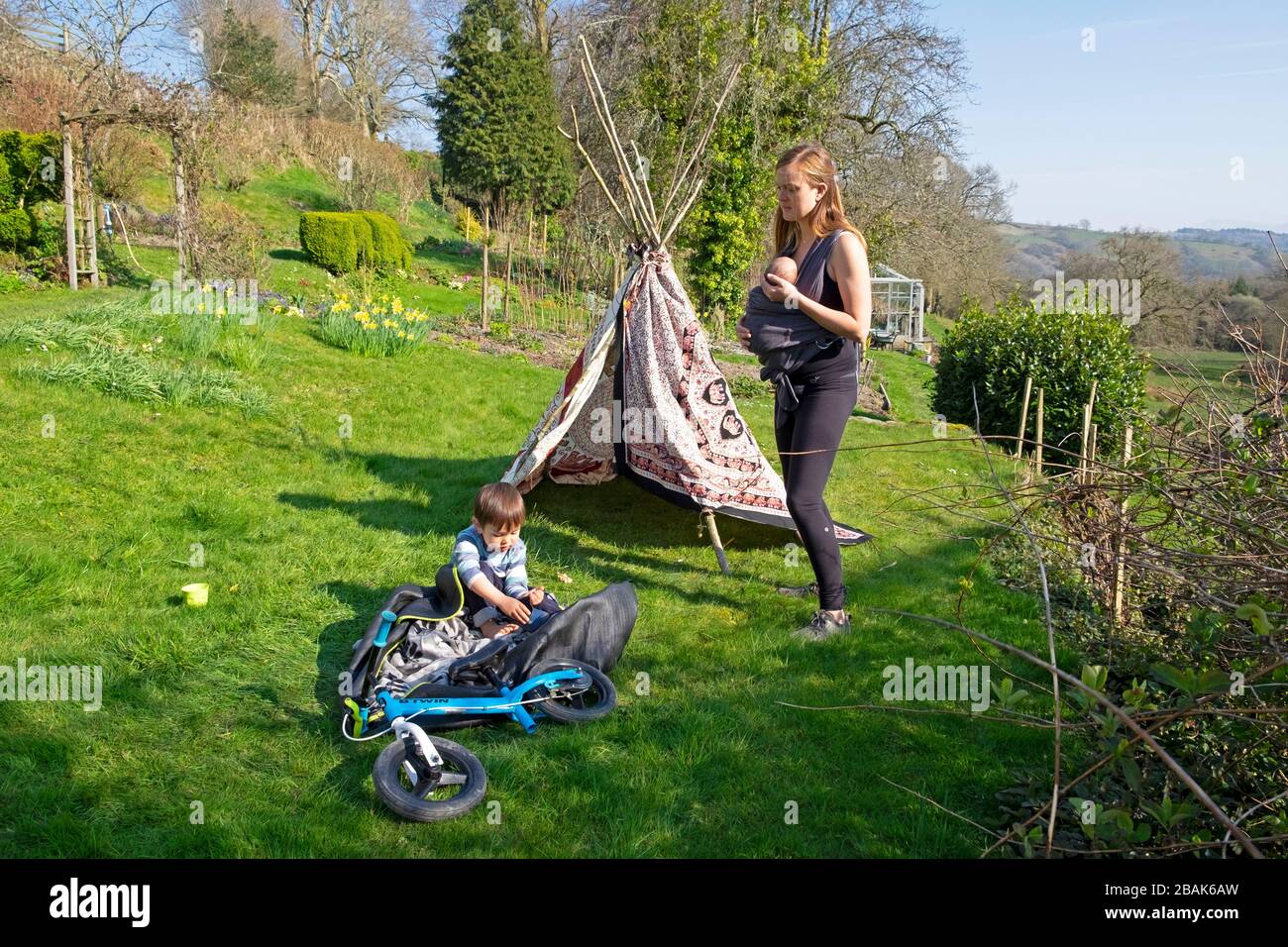 Mother standing with sling baby carrier and child boy children with tipi tent playing in a country garden outside in spring 2020 Wales UK KATHY DEWITT Stock Photo