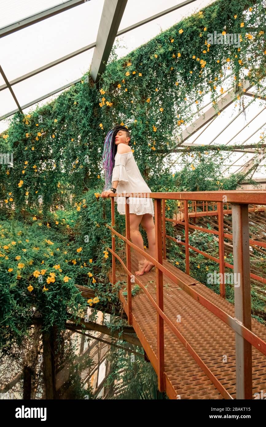 a lonely beautiful girl with multi-colored dreadlocks in a white dress stands on a red stairs in an arch of flowers. Greenhouse with exotic tropical p Stock Photo