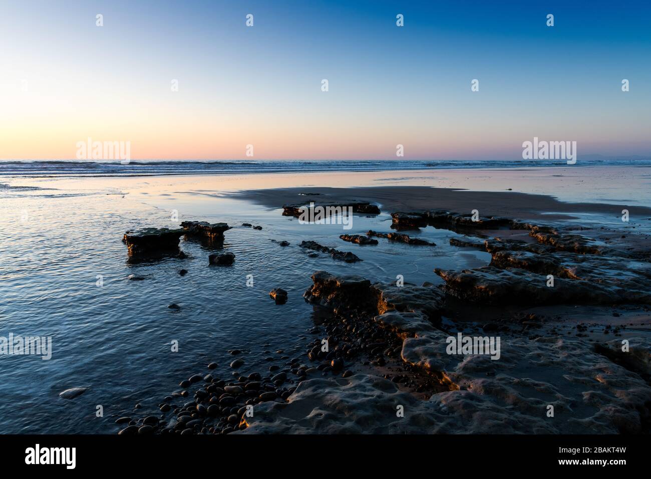 The light of a sunset casts golden highlights in pebbles and low rock formations on a beach under a purple and blue sky - Moonlight Beach, Encinitas, Stock Photo